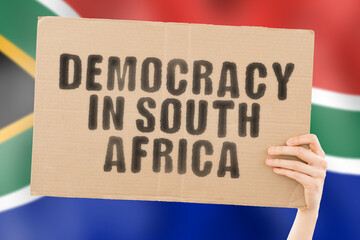 The phrase " Democracy in South Africa " on a banner in men's hand with blurred South African flag on the background. Protest. Riot. Freedom. Politics. Human rights. Liberty