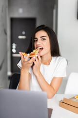 Overworked young woman taking a break and eating pizza, working from home on laptop