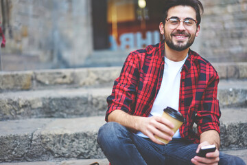 Cheerful stylish guy with smartphone sitting on stairs and enjoying coffee
