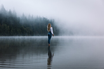 Fototapeta na wymiar Young caucasian blonde woman standing in the calm forest foggy lake.