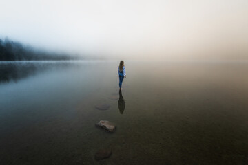 Young blonde girl standing in the foggy lake at sunrise in the wilderness forest.