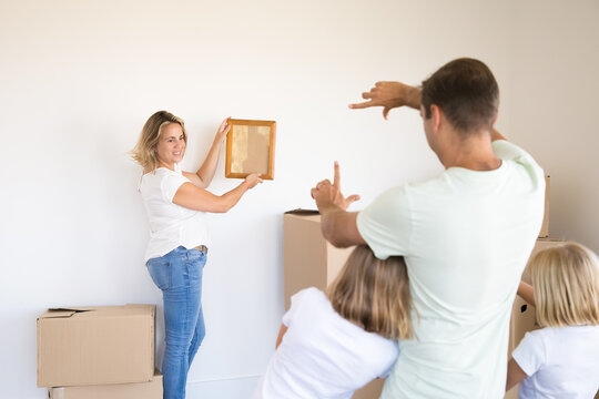 Happy family hanging picture on wall together. Pretty blonde mother moving empty frame. Father and two daughters telling her when it hanging straight. Property, relocation and moving day concept