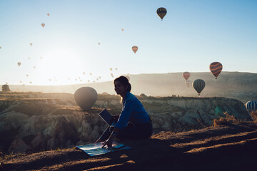 Cheerful tourist with map and laptop against hot air balloons in evening