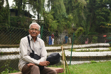 Portrait of elderly man sitting alone on bench at Lady Hydari Park in Shillong, Meghalaya, India. Happy retired old man wearing spectacles, bag, cap in hand giving pose with style looking at camera.