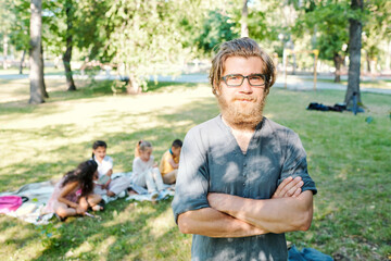 Naklejka na ściany i meble Portrait of content hipster school teacher in glasses standing with crossed arms against pupils while conducting class in park