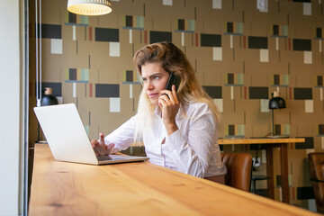 Pensive female entrepreneur using laptop and talking on mobile phone in co-working space. Young...