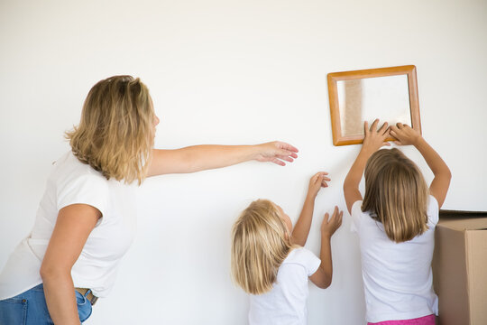 Little girl putting photo frame on white wall. Two cute daughters hanging blank picture and blonde mother helping them. Children decorating room with parent. Family, relocation and moving day concept