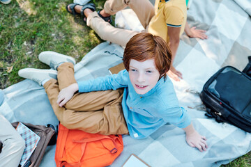 High angle view of smiling redhead schoolboy in blue shirt sitting with satchel on blanket and spending time with friends at picnic after school