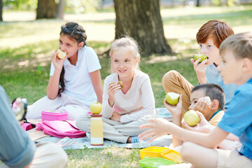 Group of positive kids sitting on plaid and eating apples while listening to teacher at outdoor...