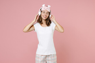 Obraz na płótnie Canvas Smiling young woman in pajamas home wear sleep mask listening music with headphones keeping eyes closed resting at home isolated on pink background studio portrait. Relax good mood lifestyle concept.