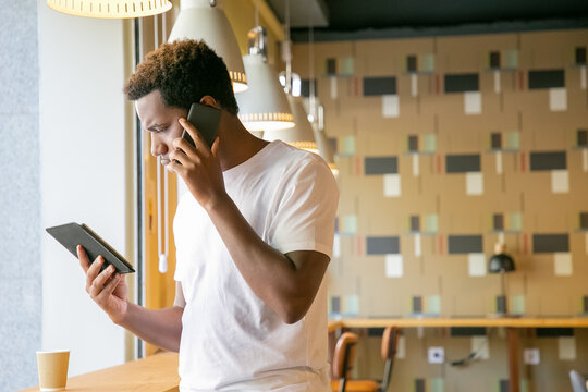 Focused African American guy talking on cell and looking at tablet screen. Young man using gadgets in co-working space or coffee shop. Wireless technology concept - Powered by Adobe