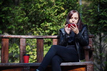 Young woman in leather jacket sitting on bench and playing on smartphone in park. Adult brunette rests among green trees in city park and browsing mobile phone.