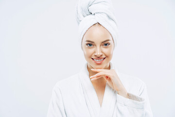 Women, beauty, hygiene, wellness concept. Fresh young European woman wears bath towel and robe, touches chin gently, spends free time in spa, undergoes beauty treatments after shower white background.