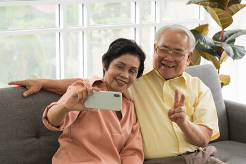 Senior Asian elderly couple in home casual outfit with happy smiling emotion sitting in living room using smartphone to take selfies with together