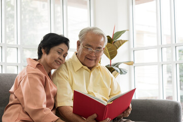 Senior Asian elderly couple in home casual outfit with happy smiling emotion sitting in living room reading photo book and bring back to the past in memory together