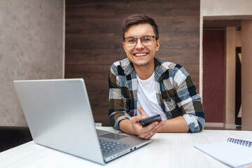 Portrait of happy young man sitting at table at home who smiles while looking at camera during break from work