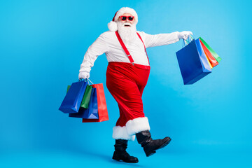 Full length profile side photo of crazy modern white grey hair bearded santa claus go shopping buy x-mas christmas bags wear red suspenders overalls boots isolated blue color background
