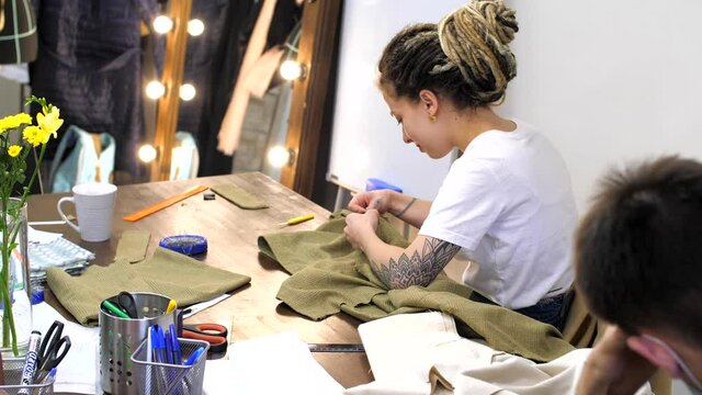 Dressmaker or seamstress working in her workshop studio at table. Fashion designer sewing new model of clothes. Adult woman with dreadlocks and tattoos sews clothes in atelier. Professional tailor