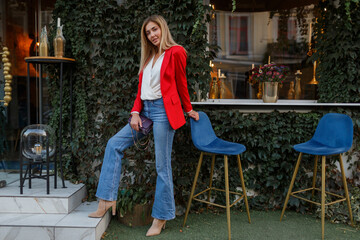 Young european  confident woman with   candid smile posing outdoor in the bar. Wearing red fashionable jacket.Traveling female enjoing vacations  in  the city.  Full lenght.