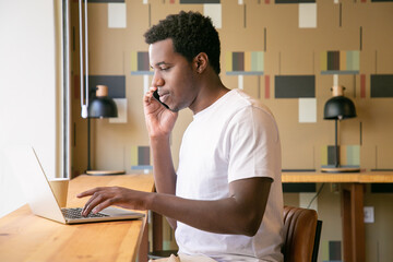 Focused young entrepreneur working at laptop and talking on mobile phone in co-working space. African American guy sitting at desk with computer and coffee. Medium shot. Communication concept