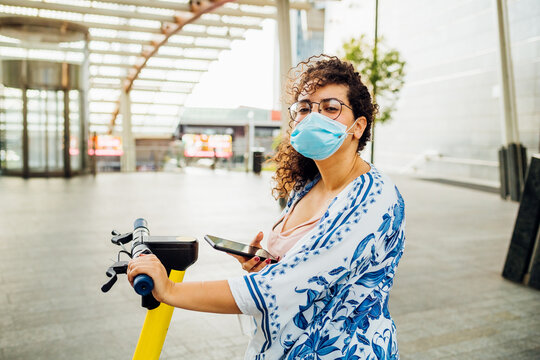 Young Multiethnic Woman Wearing Mask Unlocking Escooter - Diverse Ecological Female Outdoor Using Smartphone Driving Kickscooter