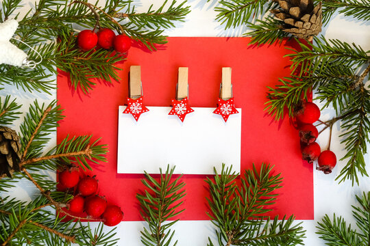 White business card with clothespins on a red photo with free space for text. Green branches of a Christmas tree, cones, berries.Christmas card, banner, letter.Happy New Year!Merry Christmas.Top view