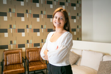 Happy successful young business woman standing with arms folded and posing in co-working or coffee shop interior, looking at camera and smiling. Medium shot. Professional portrait concept