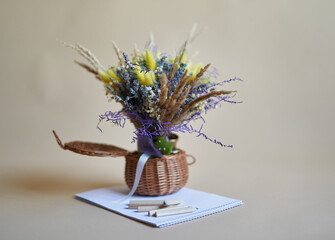 Dried flowers bouquet in wicker basket with pencils and paper near it on beige background. Knowledge Day concept.