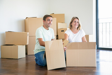 Happy couple unpacking things in new apartment, sitting on floor with open boxes, looking away. Relocation or moving concept