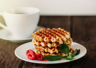 closeup view of delicious belgian waffles with raspberries and mint leaves on wooden table, cup of coffee on background 