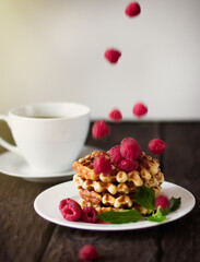 closeup view of delicious belgian waffles with raspberries and mint leaves on wooden table, cup of coffee on background 