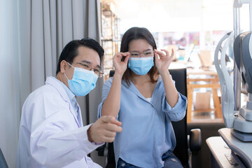 An Asian optometrist wears a protective mask and suggests a woman to look at something, women are testing new glasses by looking