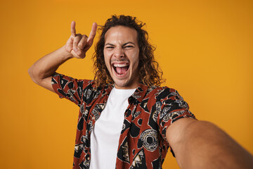 Image of excited young man taking selfie photo and showing horn gesture