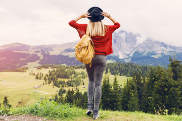 Woman  blonde traveler hipster with backpack holding hat and looking forward at amazing mountains and valley view. Space for text. Wearing stylish boho fall outfit, red knitted sweater and jeans.