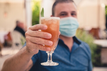 Man wearing a mask lording a drink in an  outdoor bar area 