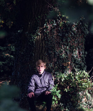 Pre-teen boy sitting in a pool of sunlight under a large oak tree.