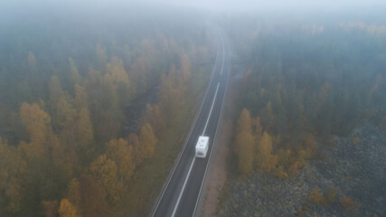 White car driving along foggy country road leading through misty forest. Atumn highway in Lapland.
