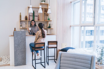 Cheerful girlfriends communicating while sitting at counter with cups of hot drink at home