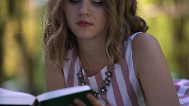 Fashionable lady with loose curly fair hair reads blurred book of blue colour lying on grass in park against green trees on sunny summer day, slow motion