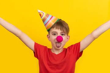 Cheerful child with red clown nose and festive hat on yellow background. April fools day.