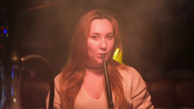 Pretty sexy woman in a beige sweater with deep neckline resting in a bar and smokes a hookah. Media. Portrait of a young long haired girl holding mouthpiece hookah by her sensitive lips.