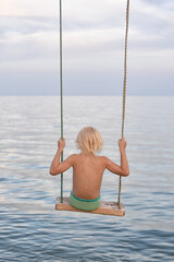 Fair-haired boy riding a rope swing over the sea. Summer holidays with children