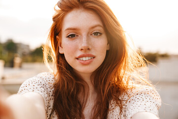 Image of pleased ginger girl taking selfie and smiling at camera