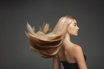 Beautiful model with long smooth, flying blonde hair isolated on dark grey studio background. Young caucasian model with well-kept skin and hair blowing on air. Concept of salon care, beauty, fashion.
