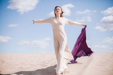 A young, slender girl in a beige dress with purple cloth in her hands posing in the desert in the wind