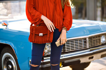 Close up fashion details of woman posing in front of vintage car, modern boho stylish trendy outfit, navy denim pants, orange blouse and bag, matching accessories, holding smartphone, spring summer.