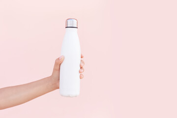 Close-up of female hand holding reusable, steel eco thermo water bottle of white, isolated on background of pastel pink color with copy space. Be plastic free. Zero waste. Environment concept.