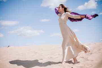 A young, slender girl in a beige dress with purple cloth in her hands posing in the desert in the wind