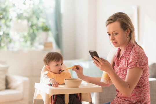 Mother feeding little boy (2-3 years) in high chair and texting message