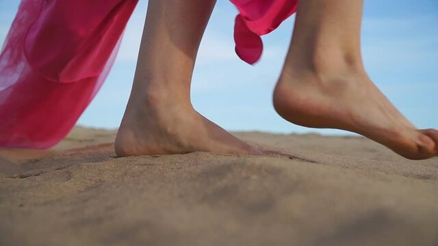 Low side view on slim female feet going along beach. Unrecognizable woman wearing red dress slowly stepping at sand enjoying resting on resort. Young barefoot girl walking in desert. Slow motion